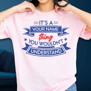 May include: A light pink t-shirt featuring blue and red text that reads: IT'S A YOUR NAME thing YOU WOULDN'T UNDERSTAND. The design includes blue banners, stars, and horizontal lines, with the word thing written in a red script font.