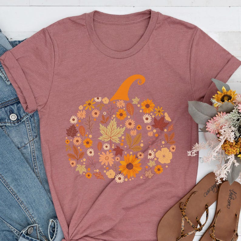 May include: Mauve-colored t-shirt featuring a floral pumpkin design. The pumpkin is composed of various flowers and leaves in shades of orange, yellow, and brown, with an orange stem. The shirt is a crew neck and has rolled-up sleeves.