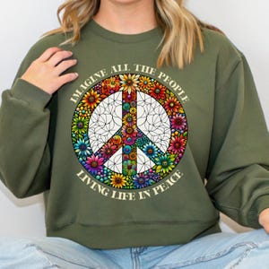 May include: Olive green sweatshirt featuring a peace sign design. The peace sign is filled with colorful flowers and surrounded by the words "IMAGINE ALL THE PEOPLE LIVING LIFE IN PEACE" in a circular arrangement.