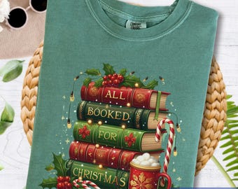 May include: Light green t-shirt with a stack of books that read "All Booked For Christmas." The books are red and green with holly and lights. A red mug with a snowflake design and candy canes are also featured.
