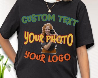May include: A black short-sleeved t-shirt featuring the text CUSTOM TEXT in blue, YOUR PHOTO in yellow, and YOUR LOGO in orange. Centered between the text is a graphic of a person with red hair holding a black camera to their face.