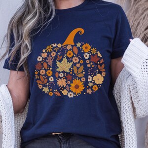 May include: Navy blue t-shirt featuring a pumpkin design made of autumn leaves and flowers in shades of orange, yellow, and brown. The pumpkin has an orange stem. A fall-themed graphic tee.