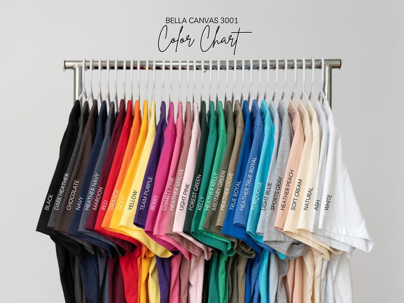 May include: A Bella Canvas 3001 color chart displaying a row of short-sleeve t-shirts hanging on white hangers. The shirts are arranged in a gradient of colors including black, dark heather, chocolate, navy, maroon, red, orange, gold, yellow, purple, pink, green, blue, gray, peach, cream, and white.