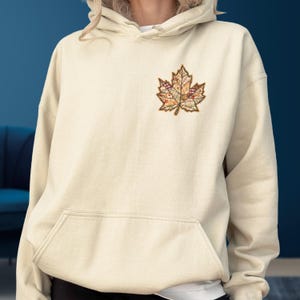 May include: A cream-colored hooded sweatshirt featuring a large embroidered maple leaf on the left chest. The leaf design is composed of various patchwork-style patterns in shades of brown, orange, yellow, and cream, with a dark brown stitched outline.