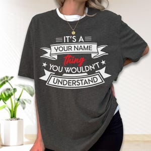 May include: A dark charcoal gray short-sleeve t-shirt featuring white and red graphic text that reads IT'S A YOUR NAME thing YOU WOULDN'T UNDERSTAND, arranged within decorative banner elements and stars on the front of the garment.