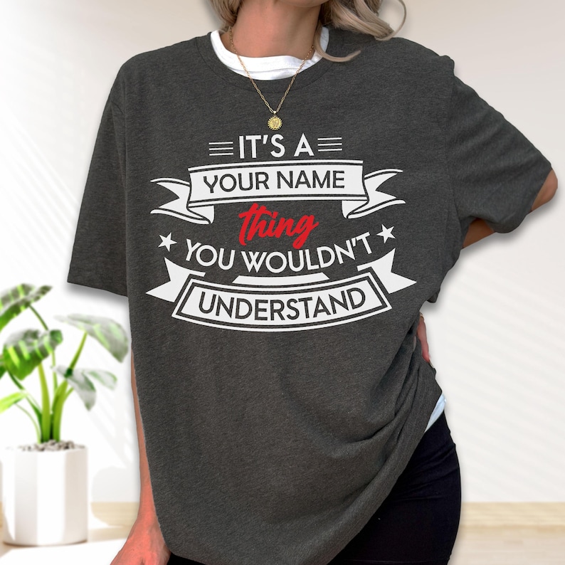 May include: A dark charcoal gray short-sleeve t-shirt featuring white and red graphic text that reads IT'S A YOUR NAME thing YOU WOULDN'T UNDERSTAND, arranged within decorative banner elements and stars on the front of the garment.