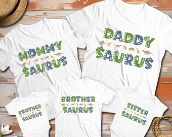 May include: A collection of white t-shirts and baby bodysuits featuring dinosaur-themed graphics. The shirts display the text Mommy Saurus, Daddy Saurus, Brother Saurus, and Sister Saurus in green and blue lettering, accented with small dinosaur illustrations.