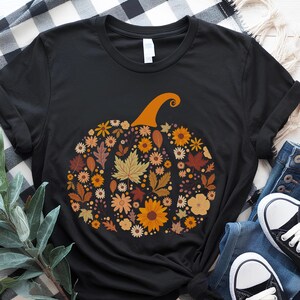 May include: Black t-shirt featuring a pumpkin design made of fall leaves and flowers in shades of orange, yellow, and brown. The pumpkin has an orange stem. The shirt is paired with blue jeans and black and white sneakers.