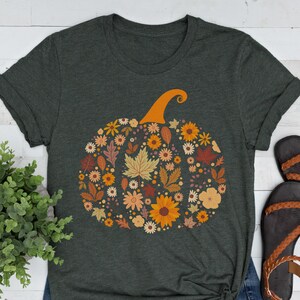 May include: Dark green t-shirt featuring a floral pumpkin design. The pumpkin is composed of various autumn-colored flowers and leaves, with an orange stem. The shirt has rolled-up sleeves and a crew neck.