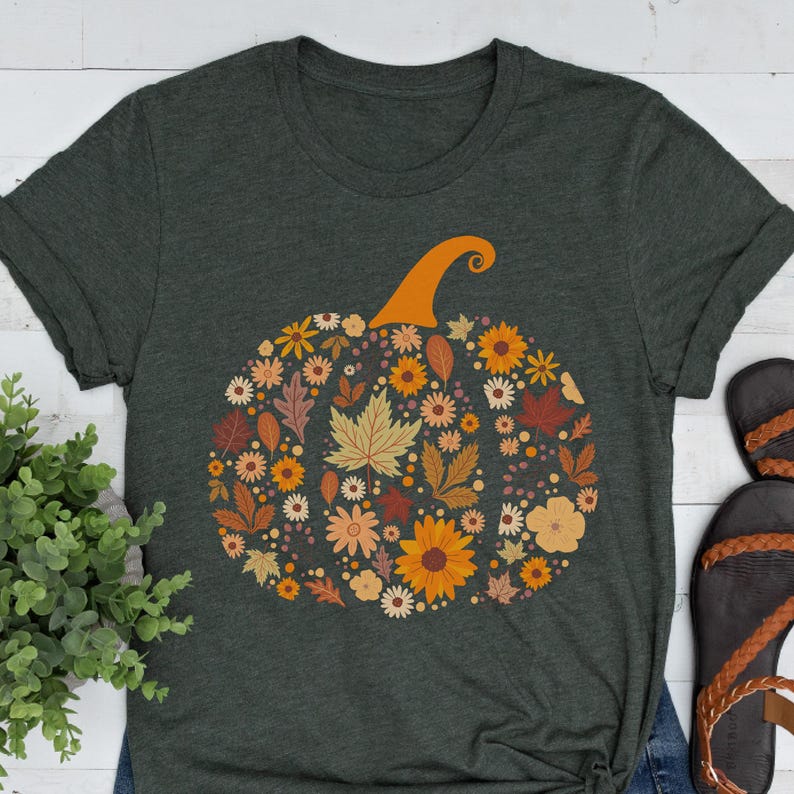 May include: Dark green t-shirt featuring a floral pumpkin design. The pumpkin is composed of various autumn-colored flowers and leaves, with an orange stem. The shirt has rolled-up sleeves and a crew neck.