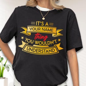 May include: A black short-sleeved t-shirt featuring yellow and red graphic text that reads IT'S A YOUR NAME thing YOU WOULDN'T UNDERSTAND, arranged within decorative yellow banners and accented with small yellow stars on the front chest area.