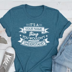 May include: A teal short-sleeved crew neck t-shirt featuring white screen-printed text that reads IT'S A YOUR NAME thing YOU WOULDN'T UNDERSTAND, arranged with decorative banners and stars, accompanied by a gray sneaker and blue denim jeans.