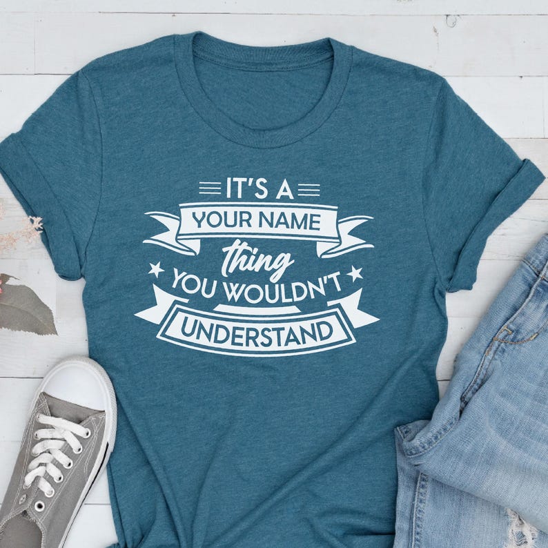 May include: A teal short-sleeved crew neck t-shirt featuring white screen-printed text that reads IT'S A YOUR NAME thing YOU WOULDN'T UNDERSTAND, arranged with decorative banners and stars, accompanied by a gray sneaker and blue denim jeans.