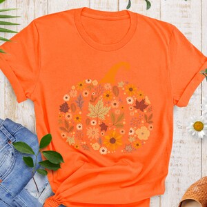 May include: Bright orange t-shirt featuring a floral pumpkin design. The pumpkin is composed of various flowers and leaves in shades of yellow, orange, and brown. The shirt has short sleeves and a crew neck.