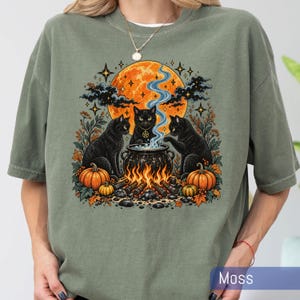 May include: A moss green t-shirt featuring a graphic print of three black cats gathered around a bubbling cauldron over a fire. The scene includes a large orange moon, autumn leaves, pumpkins, and stars. The word Moss is printed in the corner.