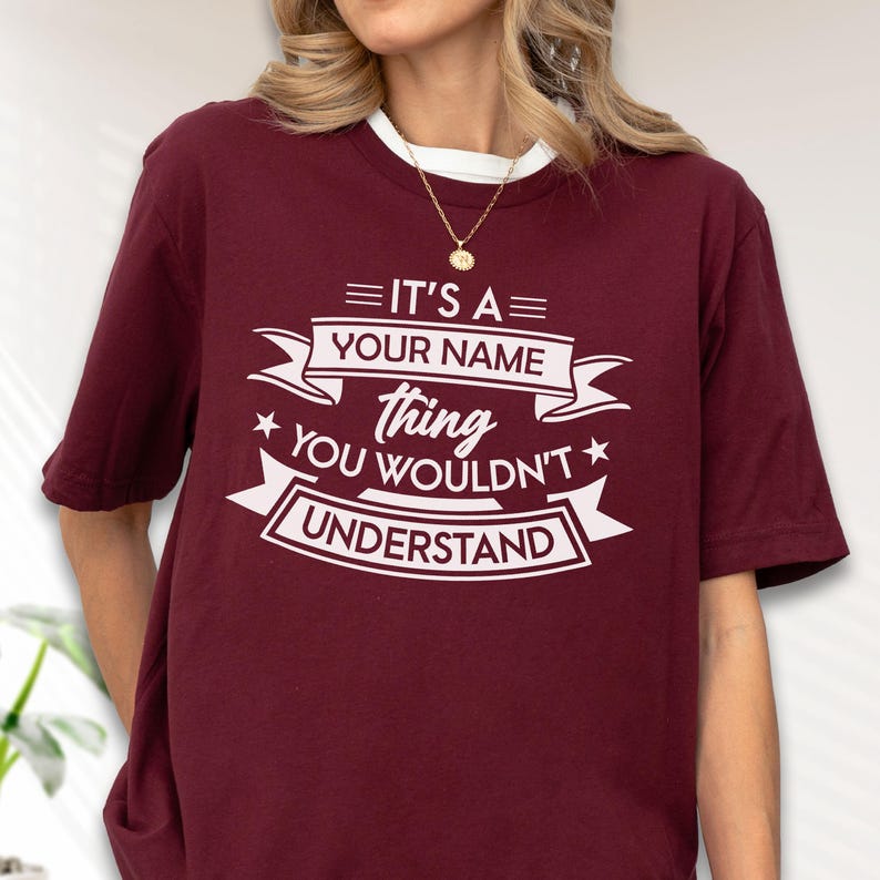 May include: A maroon short-sleeved t-shirt featuring white screen-printed text that reads: IT'S A YOUR NAME thing YOU WOULDN'T UNDERSTAND. The design includes decorative banners and stars, centered on the chest of the crew neck garment.