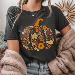 May include: Dark gray t-shirt featuring a floral pumpkin design. The pumpkin is filled with various autumn-colored flowers and leaves, including sunflowers, maple leaves, and daisies. The pumpkin has an orange stem. The shirt is a casual, short-sleeved style.