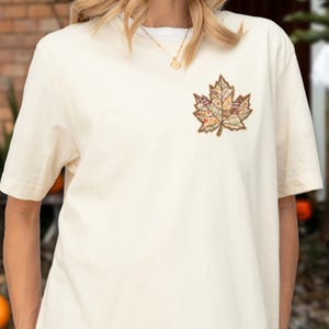 May include: Cream-colored short-sleeve t-shirt featuring a detailed maple leaf applique. The leaf design incorporates various patterned fabrics in shades of brown, orange, and green, creating a textured, autumnal aesthetic. The shirt has a classic crew neck.