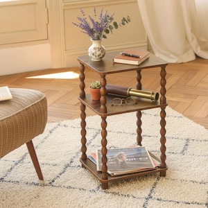 May include: A three-tiered, dark brown wooden side table with turned legs. The top shelf holds a vase of purple flowers, a book, and a small succulent. The middle shelf has a telescope and glasses. The bottom shelf has magazines.