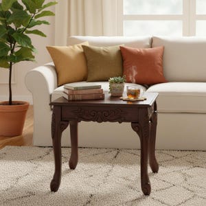 May include: A dark brown, ornate side table with curved legs and a carved apron. The table holds books, a small potted plant, and a cup of tea. Three throw pillows in shades of gold, green, and orange sit on a white sofa in the background.