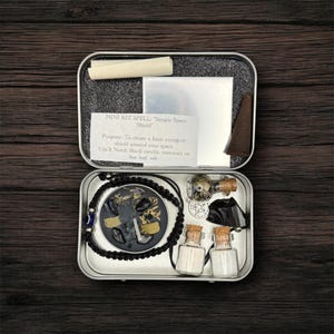 May include: An open metal tin containing a mini spell kit. The kit includes a bracelet, small glass bottles, a cone, and a paper with text that reads "MINI KIT SPELL: Simple Space Shield". The kit is on a dark wooden surface.
