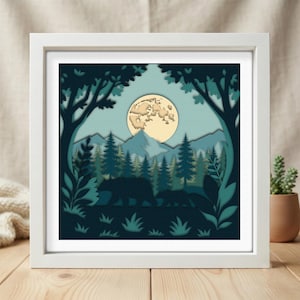 May include: A square white frame containing a layered paper art piece depicting a forest scene at night. The artwork features silhouettes of two bears walking through a pine forest with mountains in the background under a large, pale yellow moon.