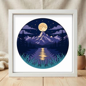 May include: A square white frame containing a circular layered paper art piece depicting a night landscape. The scene features a pale yellow full moon above purple mountains, dark blue pine trees, a reflective lake, and teal grass in the foreground.