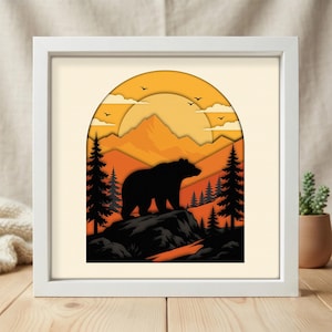 May include: A square white frame containing a layered paper art piece depicting a black bear silhouette standing on a rock. The scene features orange and yellow mountains, pine trees, and a large sun in the background with small birds flying.