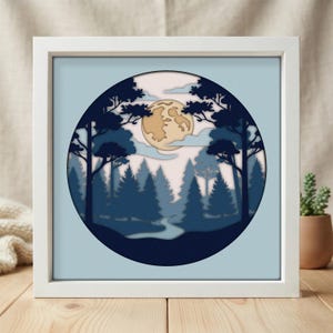 May include: A square white frame containing a circular layered paper art piece depicting a forest scene at night. The design features dark blue silhouettes of pine trees, a winding light blue path, pale clouds, and a large beige full moon.