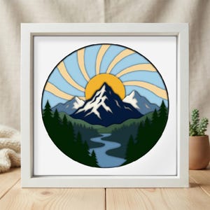 May include: A square white frame containing a circular layered paper art piece. The design features a dark blue mountain range with white snow-capped peaks, a winding blue river, dark green pine trees, and a yellow sun with light blue rays.