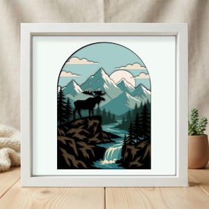 May include: A square white frame containing a layered paper art piece with an arched cutout. The scene depicts a black silhouette of a moose standing on a rocky cliff overlooking a river, a waterfall, pine trees, and snow-capped mountains.