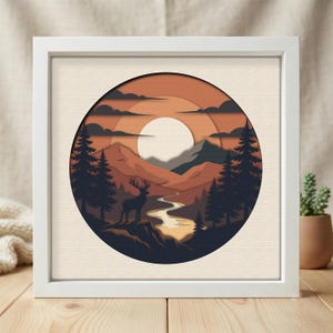 Puede incluir: Un marco cuadrado blanco que contiene una obra de arte circular de papel en capas. La imagen muestra la silueta de un ciervo sobre un acantilado frente a un río sinuoso, pinos, montañas y un gran sol en un cielo naranja con nubes.