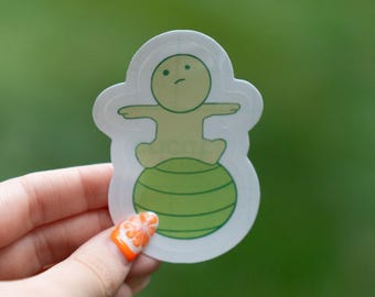 May include: A sticker featuring a cartoon figure with arms outstretched, seated on a green exercise ball. The figure and ball are outlined in white, with a green background. The sticker is held by a hand with an orange and white nail design.