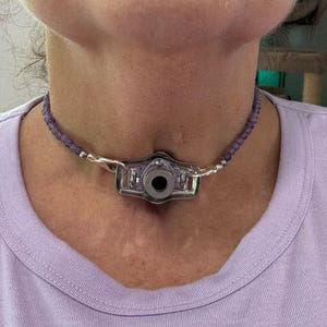 以下が含まれることがあります： 黒い円形要素を備えた中央のペンダントが付いたシルバーカラーのネックレス。ネックレスは小さな紫色のビーズで飾られています。ペンダントは、シルバーのリンクでビーズストランドに接続されています。ネックレスは首の周りに着用されています。