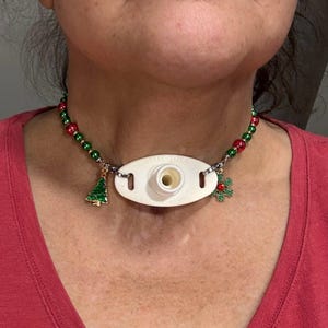 May include: A Christmas-themed necklace with red and green beads. The necklace features a white medical device with the text "MOORE SIZE" and charms of a green Christmas tree and a red and green holly. The necklace is worn around the neck.