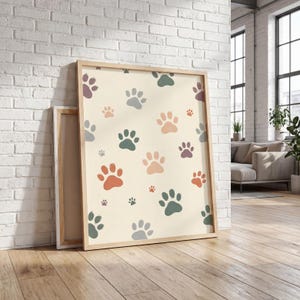May include: A framed canvas print featuring a repeating pattern of colorful paw prints in shades of orange, green, purple, and pink on a cream background. The print is displayed against a white brick wall and wooden floor.