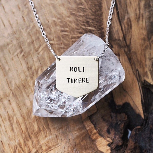 May include: A silver chain necklace with a brass pendant that reads "NOLI TIMERE". The pendant is shaped like a shield and is hanging on a clear quartz crystal.