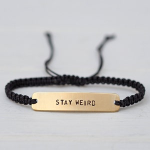 Könnte beinhalten: Schwarzes Makramee-Armband mit einer goldfarbenen rechteckigen Platte. Auf der Platte steht in serifenlosen Buchstaben "STAY WEIRD". Das Armband ist mit schwarzen Kordeln verstellbar.