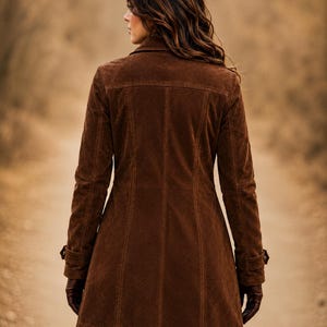 May include: A dark brown suede coat featuring a structured collar, long sleeves with buttoned cuff straps, and vertical seam detailing along the back. The coat has a mid-thigh length and a fitted silhouette, paired with dark brown leather gloves.