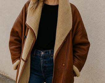 May include: A brown leather coat with a wide, beige shearling-style collar and lining. The coat features long sleeves with shearling cuffs, a toggle closure on the front, and a smooth, matte finish on the outer leather surface.