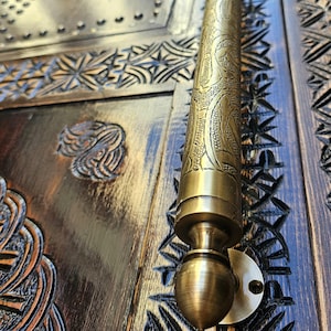 Op de afbeelding: Een sierlijke messing deurklink met ingewikkelde bloemen- en bladerpatronen. De cilindrische handgreep is gemonteerd op een donkere houten deur met gesneden geometrische ontwerpen. De deur heeft een verweerde afwerking.