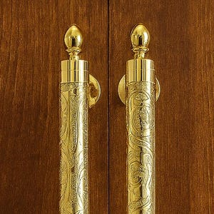 May include: Two ornate, gold-colored cabinet pulls with a detailed, swirling pattern. The pulls are cylindrical with decorative finials at the top and bottom. They are mounted on a dark brown wooden cabinet door, creating a classic look.