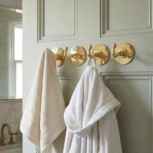 May include: A row of four brass-colored hooks mounted on a light green door. A cream-colored towel and a white hooded robe hang from the hooks. The bathroom setting includes a sink, mirror, and faucet.