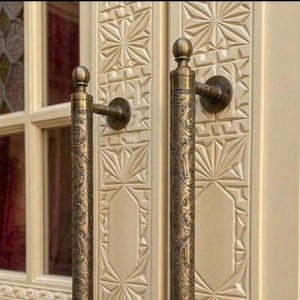 May include: Close-up of two ornate bronze door handles with intricate floral patterns. The handles are mounted on a cream-colored door with carved geometric designs. The door has a window with a stained glass design.