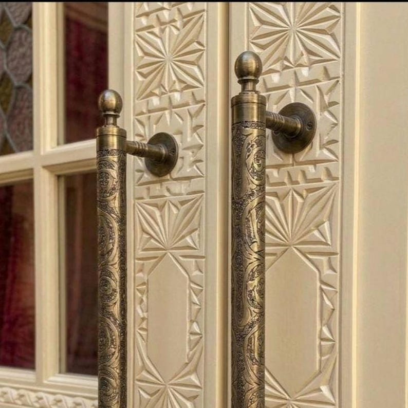 May include: Close-up of two ornate bronze door handles with intricate floral patterns. The handles are mounted on a cream-colored door with carved geometric designs. The door has a window with a stained glass design.