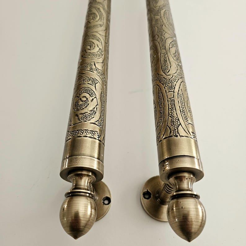 May include: Two antique bronze door handles with intricate floral and scrollwork patterns. The handles have a cylindrical shape with decorative end pieces and circular mounting plates. The metal has a warm, aged patina, suggesting a vintage or handcrafted origin.