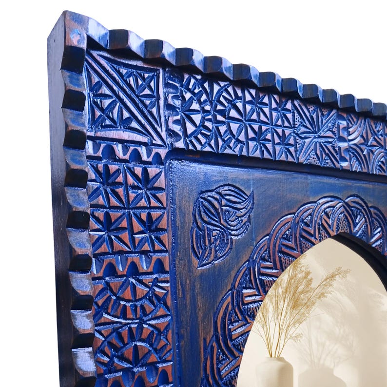 May include: Close-up of a blue wooden mirror frame with intricate carved geometric patterns. The frame has a distressed finish, revealing hints of brown underneath the blue paint. The design includes floral and star-like motifs.