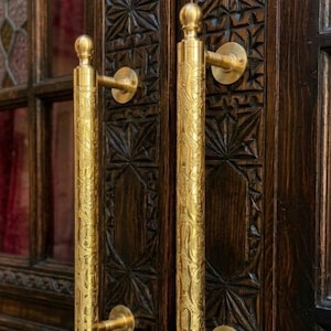 May include: Close-up of a dark wooden door with ornate carvings and two gold-colored door pulls. The pulls are cylindrical with decorative patterns and a rounded top. The door has a vintage aesthetic.