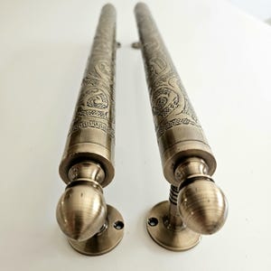 May include: Two antique bronze door handles. Each handle features an ornate, engraved design along its cylindrical body. The handles have a rounded base and a decorative, spherical end. The hardware is mounted on a white surface.