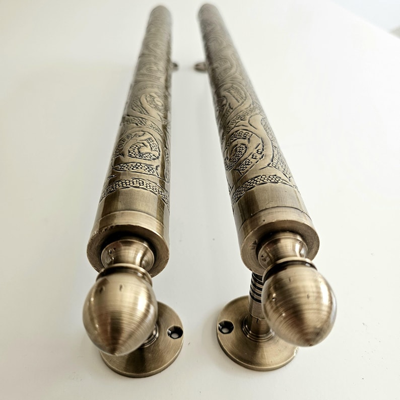 May include: Two antique bronze door handles. Each handle features an ornate, engraved design along its cylindrical body. The handles have a rounded base and a decorative, spherical end. The hardware is mounted on a white surface.